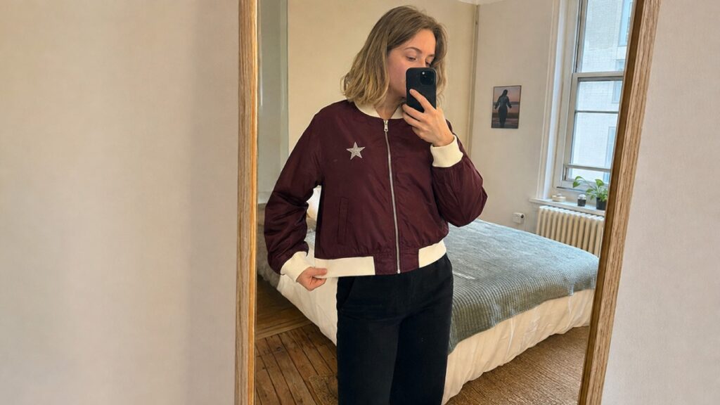 A phone mirror selfie of someone wearing a burgundy bomber jacket with a cream collar and cuffs and a silver star on the chest — the kind of reference photo a customer sends in.