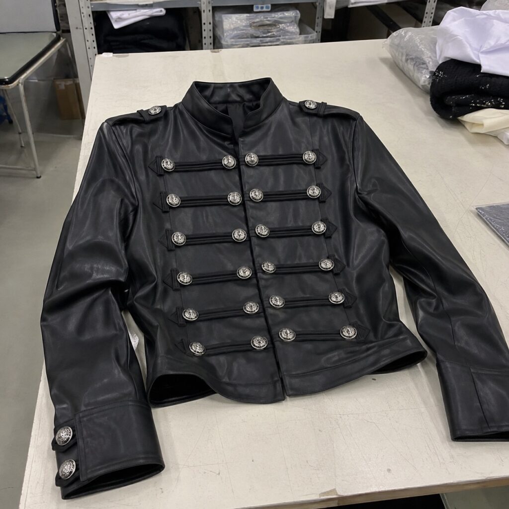 A black leather jacket with two rows of silver buttons and a stand collar, laid out on a cutting table in a factory for quality control.