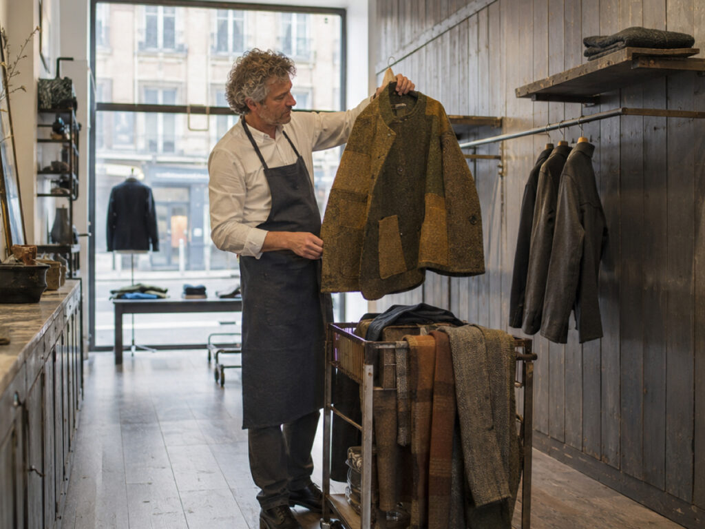 A shopkeeper in an apron holding up a jacket in a small boutique, rails of clothing either side.