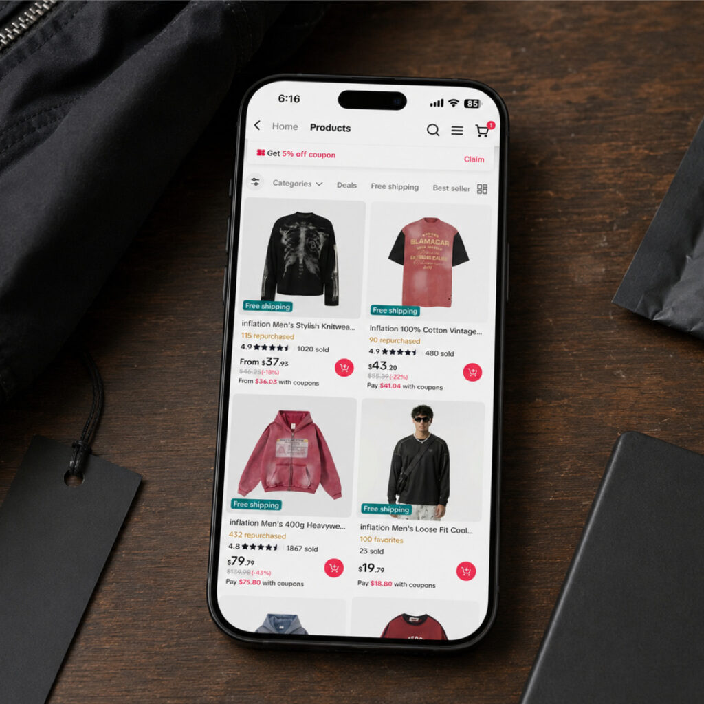 A phone showing a marketplace product grid of jackets and tops, photographed on a dark desk beside folded stock.