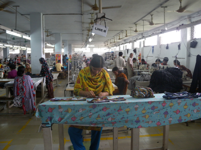 A garment factory floor with rows of sewing machines under a sign reading LINE-B, workers seated at the machines and finished pieces stacked on a table in the foreground