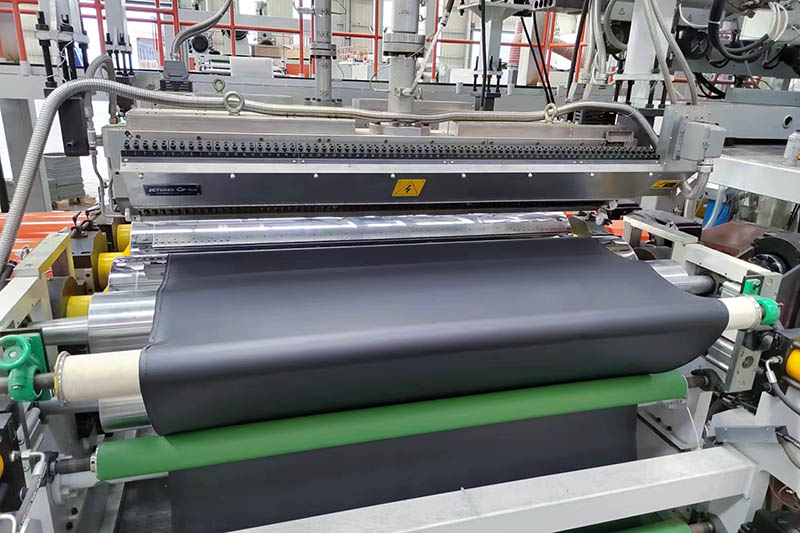 Industrial coating line applying a dark polymer coating to wide textile material through rollers.