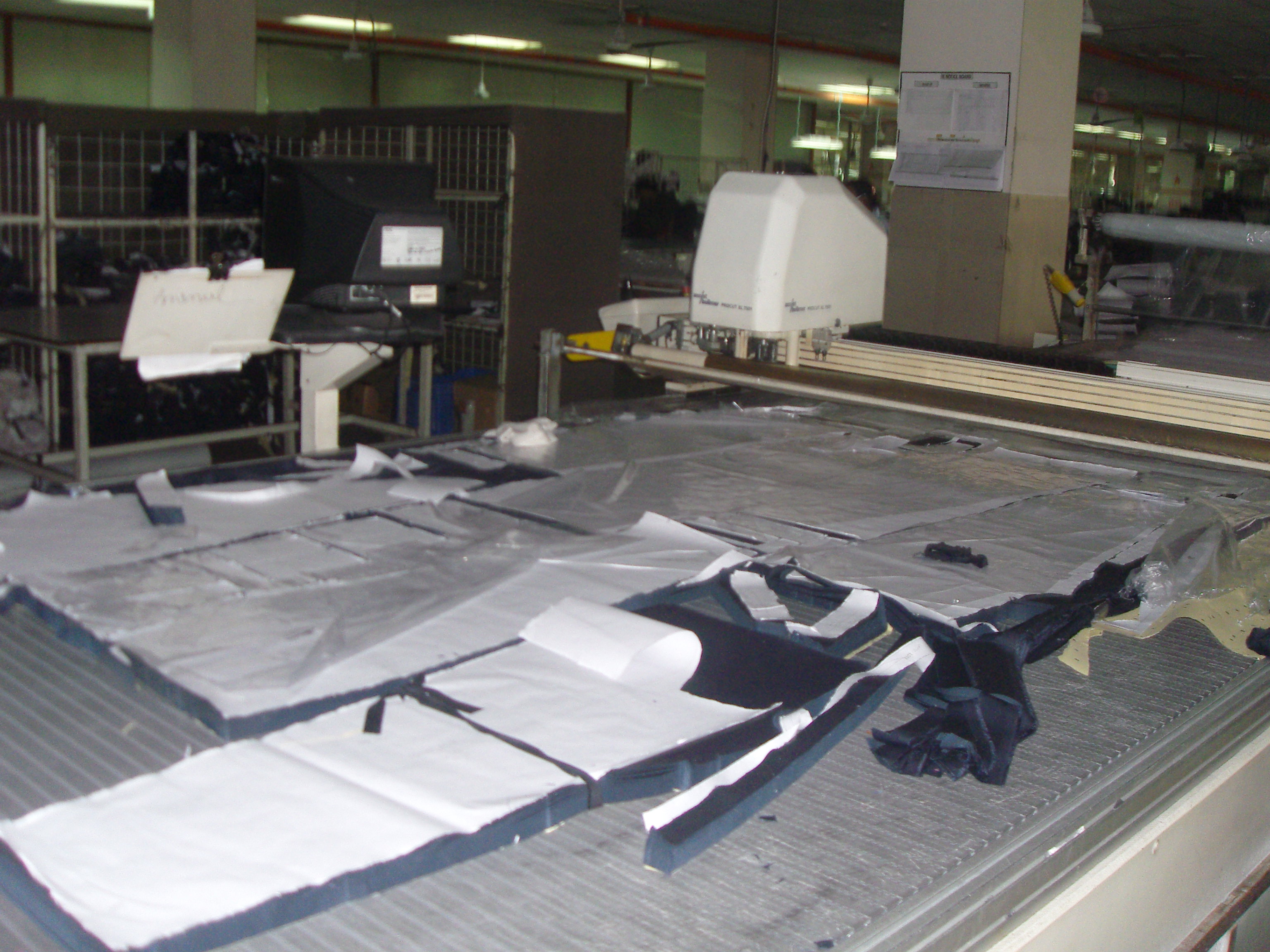 A cutting table in a garment factory with dark fabric spread in layers under plastic film, cut pattern pieces lying across the lay, and the head of an automatic cutter on its rail to the right