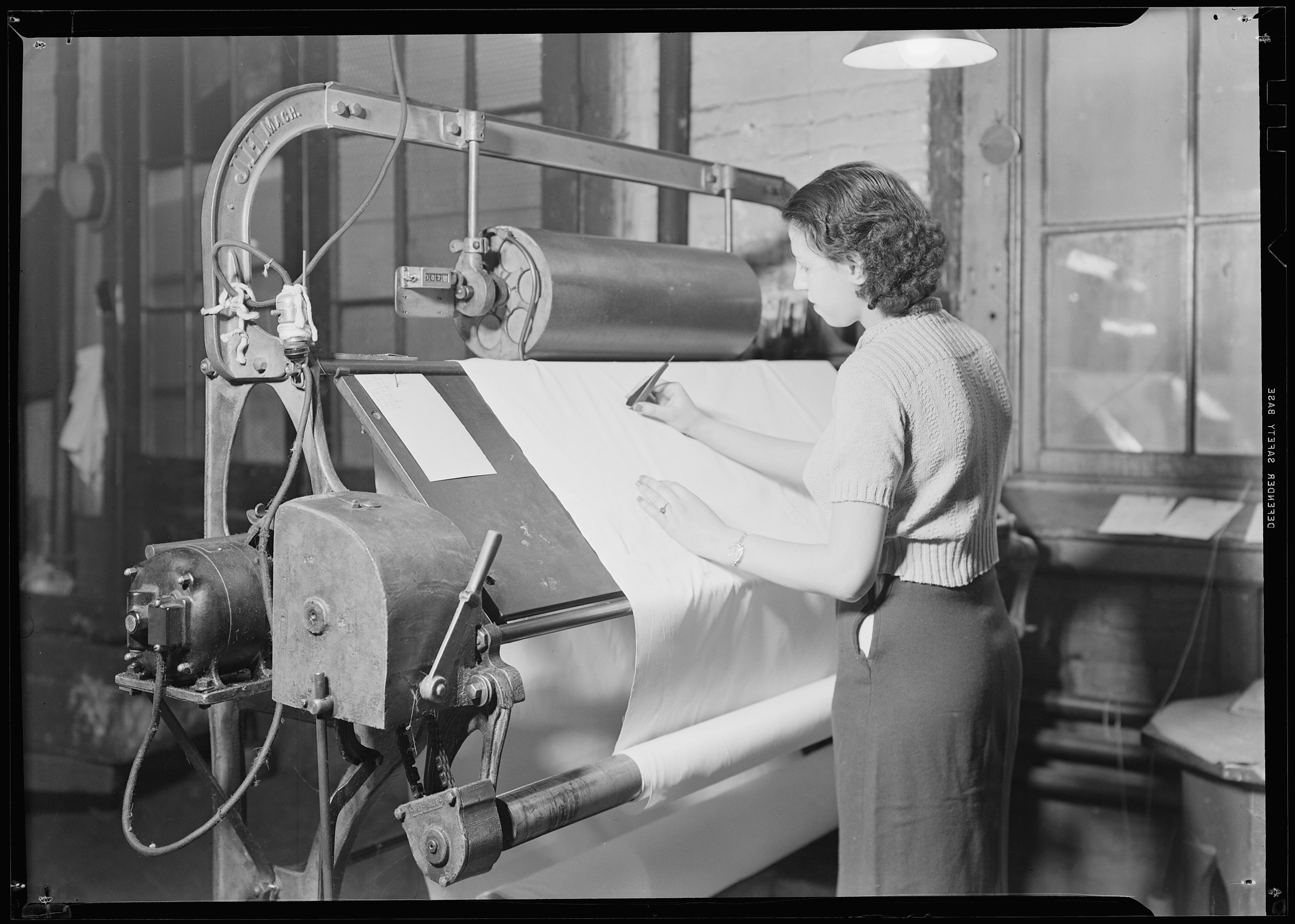 A woman at a fabric inspection frame, cloth running from a lower beam across a lit board, pencil in hand to mark defects as the roll passes