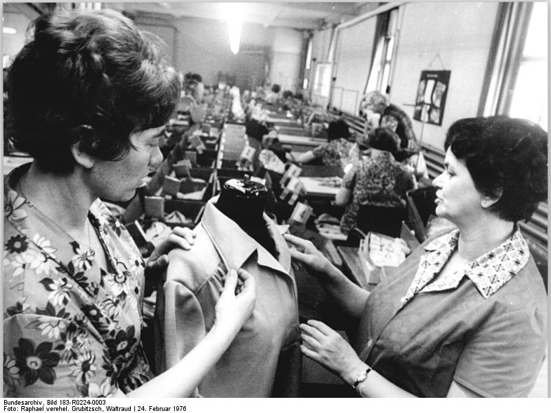 Two garment workers inspect the collar and front construction of a finished jacket on a dress form at a clothing factory